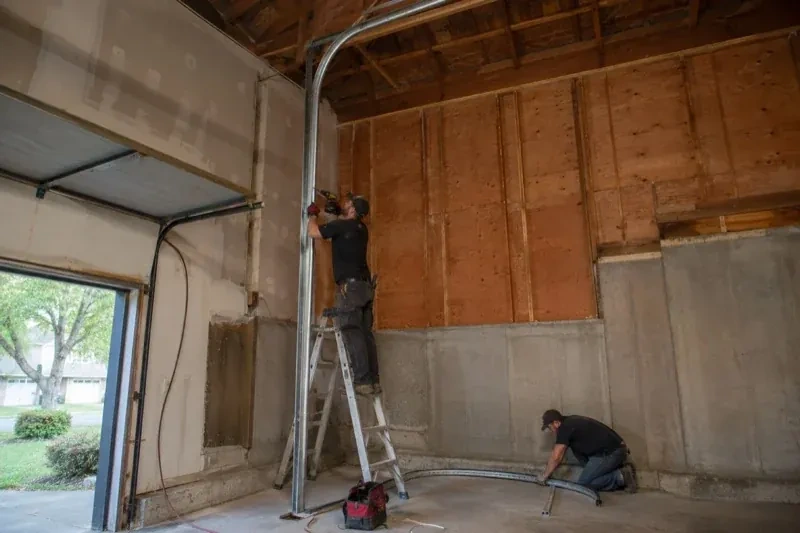 One person on a stepladder working beside a tall curved garage-door track while another kneels beside loose track sections in an unfinished garage. — High-Lift Garage Door Conversion, Waterloo